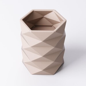 May include: A beige geometric planter with a faceted design. The planter has a hexagonal top and a cylindrical body with a repeating diamond pattern. The neutral color and modern design make it suitable for various decor styles.