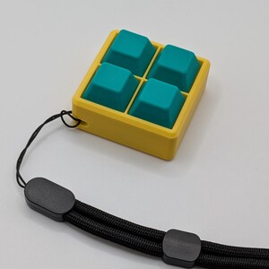 May include: A yellow square container with four teal-colored, textured square shapes inside. A black wrist strap is attached to the container. The wrist strap is made of a black woven material and has black plastic clasps.