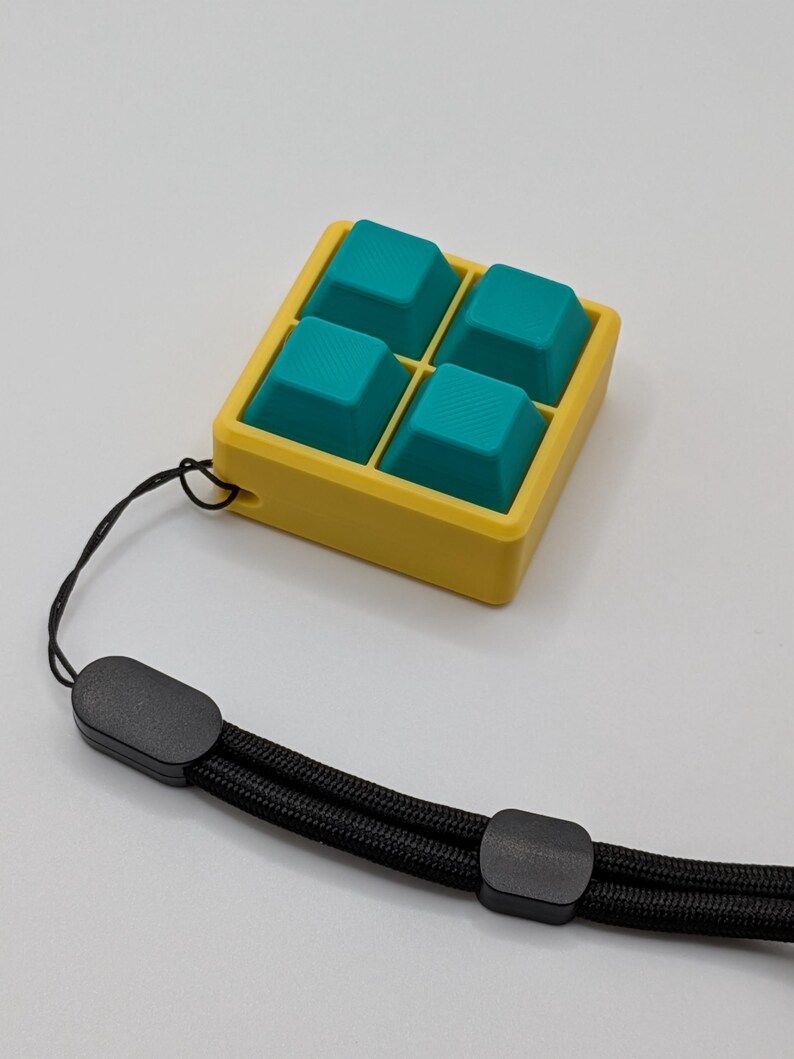 May include: A yellow square container with four teal-colored, textured square shapes inside. A black wrist strap is attached to the container. The wrist strap is made of a black woven material and has black plastic clasps.