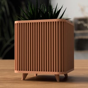 May include: A peach-coloured, square planter with vertical slat detailing. The planter has short, rounded legs and contains a green plant with spiky leaves. The planter is on a wooden surface.