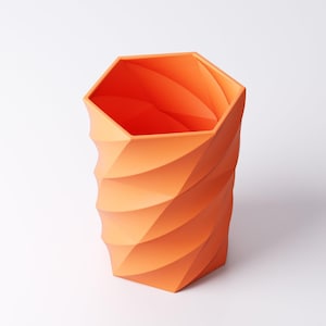 May include: An orange geometric vase with a hexagonal top and a spiraling, textured design. The vase is made of a solid material and has a modern, minimalist aesthetic. The vase is approximately 8 inches tall.