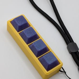 May include: A yellow rectangular case holds four square, blue-purple keycaps. A black lanyard is attached to the case. The keycaps have a glittery, textured appearance.