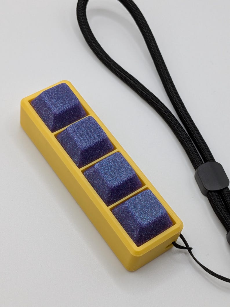 May include: A yellow rectangular case holds four square, blue-purple keycaps. A black lanyard is attached to the case. The keycaps have a glittery, textured appearance.