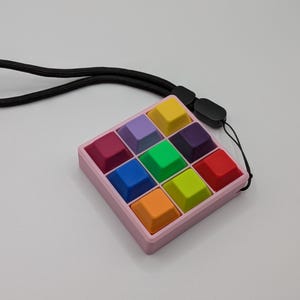 May include: A colorful, square fidget toy with a pink frame and twelve multi-colored square buttons. The buttons are yellow, purple, green, blue, orange, and red. A black lanyard is attached to the toy.