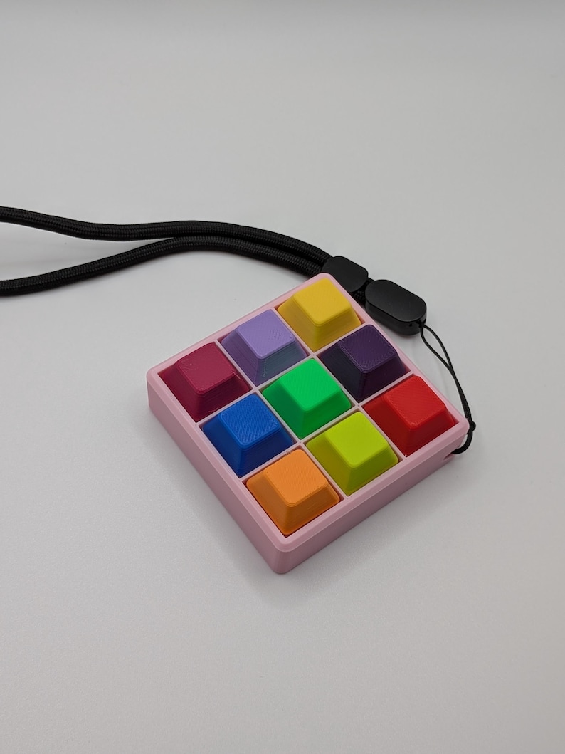 May include: A colorful, square fidget toy with a pink frame and twelve multi-colored square buttons. The buttons are yellow, purple, green, blue, orange, and red. A black lanyard is attached to the toy.