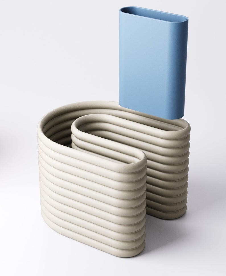 May include: An abstract sculpture comprising two beige, wave-like forms and a light blue, rectangular vase. The beige forms have a ribbed texture, while the blue vase is smooth. The artwork is set against a white backdrop.