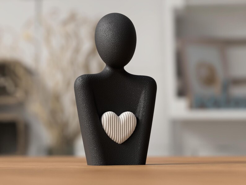 Puede incluir: Figura negra de una persona sosteniendo un coraz&oacute;n. El coraz&oacute;n es blanco con l&iacute;neas verticales. La figura tiene una superficie texturizada y est&aacute; sobre una mesa de madera.
