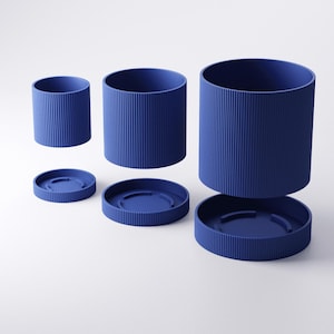 May include: A set of three blue cylindrical containers with matching lids. The containers have vertical ribbed detailing and are arranged at different heights, with one lid detached. The objects are set against a white background.