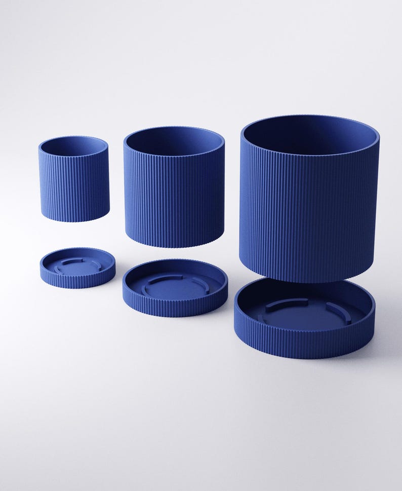 May include: A set of three blue cylindrical containers with matching lids. The containers have vertical ribbed detailing and are arranged at different heights, with one lid detached. The objects are set against a white background.