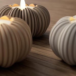 May include: Three decorative candles with a pumpkin-like design. The candles are in shades of beige and gray, with a ribbed texture. Each candle has a glowing, warm-toned flame. The candles are arranged on a wooden surface.