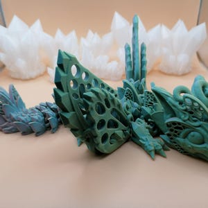 Pu&ograve; includere: Un drago articolato stampato in 3D nei toni del verde e del verde acqua, con un design cristallino dettagliato. Il drago &egrave; posizionato su una superficie neutra, con formazioni di cristalli bianchi sullo sfondo. Il corpo del drago &egrave; segmentato.