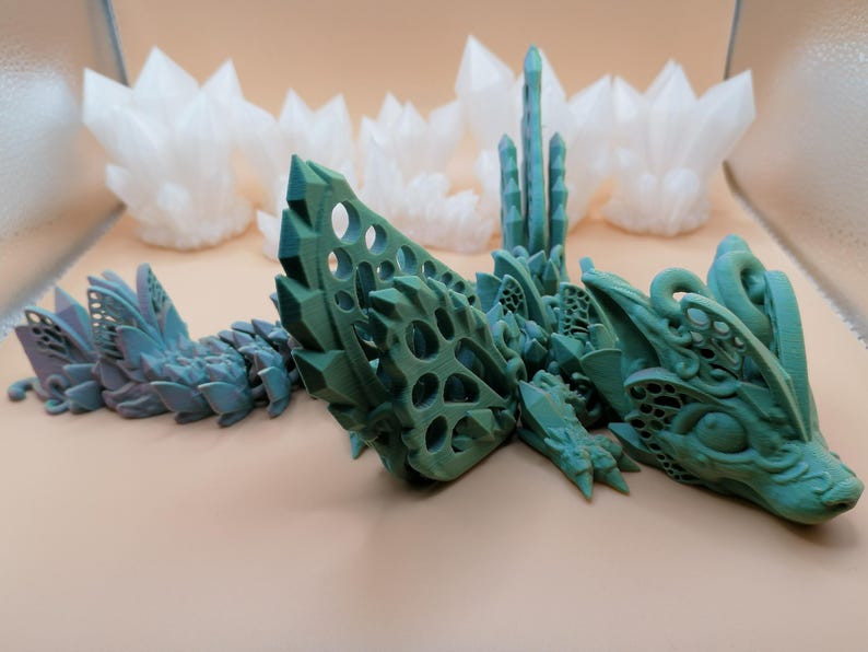 Pu&ograve; includere: Un drago articolato stampato in 3D nei toni del verde e del verde acqua, con un design cristallino dettagliato. Il drago &egrave; posizionato su una superficie neutra, con formazioni di cristalli bianchi sullo sfondo. Il corpo del drago &egrave; segmentato.