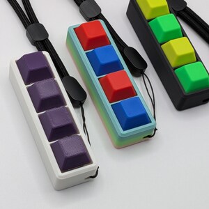 May include: Three keycap pendants with black lanyards. One pendant has purple keycaps in a white frame. Another has red and blue keycaps in a frame with a gradient of colors. The third has green and yellow keycaps in a black frame.