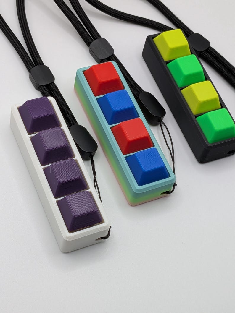 May include: Three keycap pendants with black lanyards. One pendant has purple keycaps in a white frame. Another has red and blue keycaps in a frame with a gradient of colors. The third has green and yellow keycaps in a black frame.
