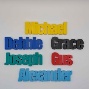 May include: A collection of colorful 3D-printed names: Michael in yellow, Debbie in blue, Grace in black, Joseph in teal, Gus in red, and Alexander in light blue. The names are displayed on a white background.
