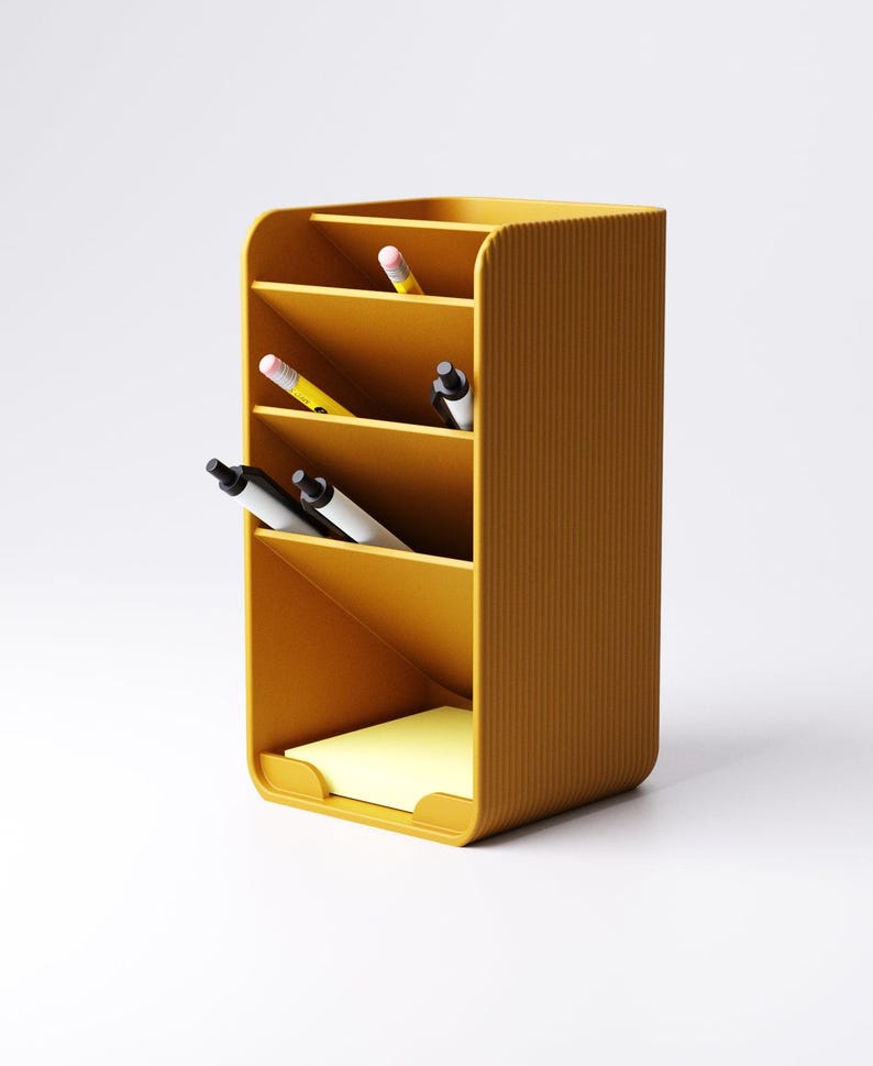 May include: Mustard yellow desk organizer with multiple compartments. The organizer holds pens, pencils, and a pad of yellow sticky notes. The exterior has a ribbed texture. The design is modern and minimalist, suitable for office or home use.