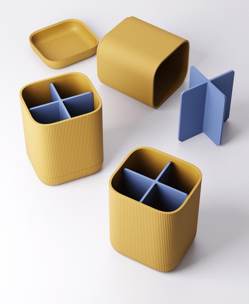 May include: A set of mustard yellow desk organizers with a ribbed texture. The set includes two square containers with blue dividers, a square lid, and a separate blue cross-shaped divider. The organizers are designed for office or home use.