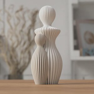 Puede incluir: Dos esculturas abstractas de madera, una grande y otra peque&ntilde;a, de pie una al lado de la otra sobre una superficie de madera. Las esculturas son de color beige claro y presentan ranuras verticales, lo que les da un aspecto texturizado. La escultura m&aacute;s grande mide unos 30 cm de altura.