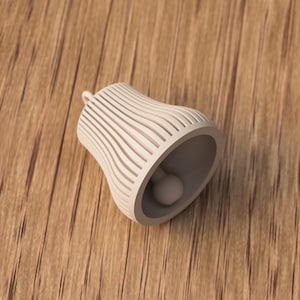 May include: A cream-colored bell-shaped ornament with a small loop at the top. The bell has a series of vertical, curved lines that create a striped effect. Inside the bell is a small, round object. The bell is lying on a wooden surface.