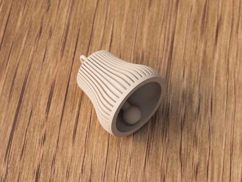 May include: A cream-colored bell-shaped ornament with a small loop at the top. The bell has a series of vertical, curved lines that create a striped effect. Inside the bell is a small, round object. The bell is lying on a wooden surface.