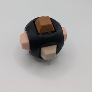 May include: A black fidget cube with six sides, each featuring a different button. The buttons are various colors, including brown, beige, and light pink. The cube is designed for stress relief and sensory stimulation.