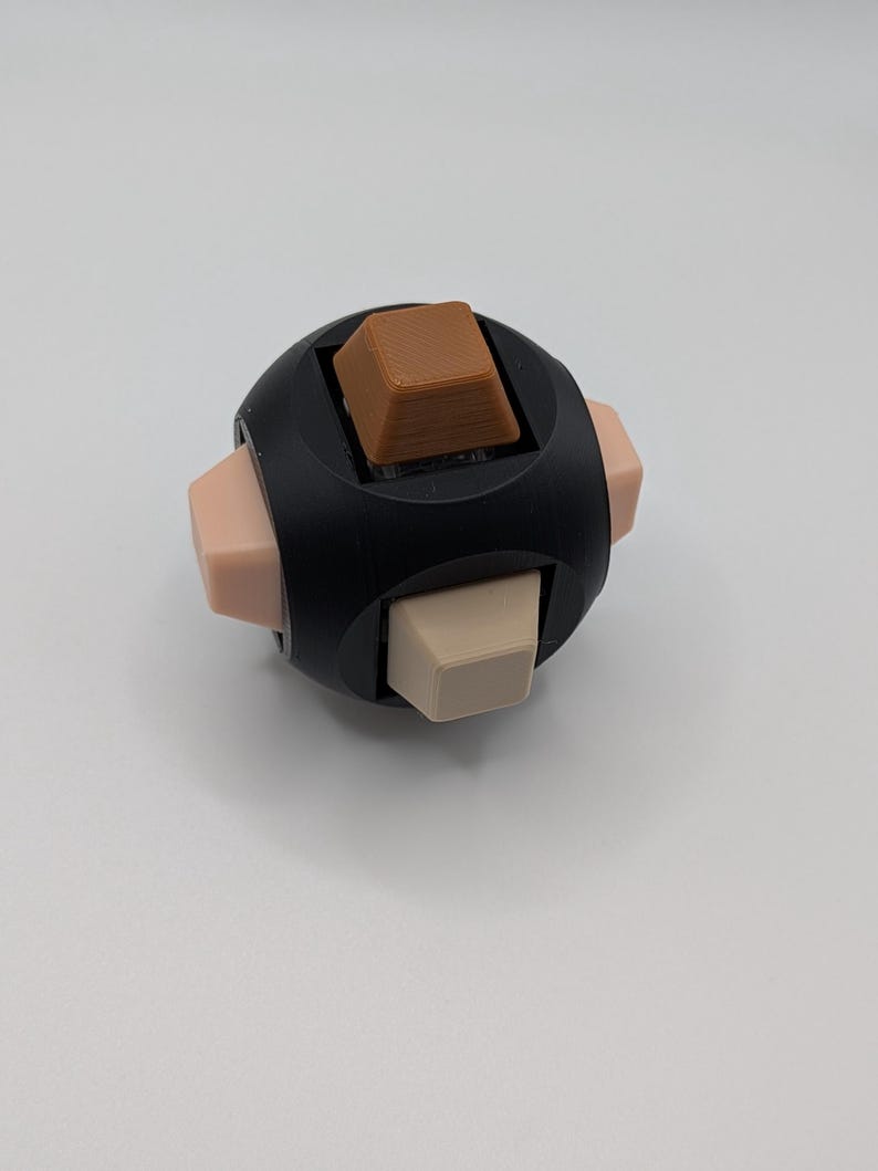 May include: A black fidget cube with six sides, each featuring a different button. The buttons are various colors, including brown, beige, and light pink. The cube is designed for stress relief and sensory stimulation.