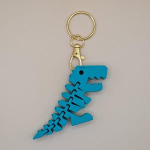 May include: A turquoise dinosaur skeleton keychain with a gold-colored clasp and keyring. The dinosaur is made of interlocking pieces, creating a flexible design. The keychain is a fun accessory.