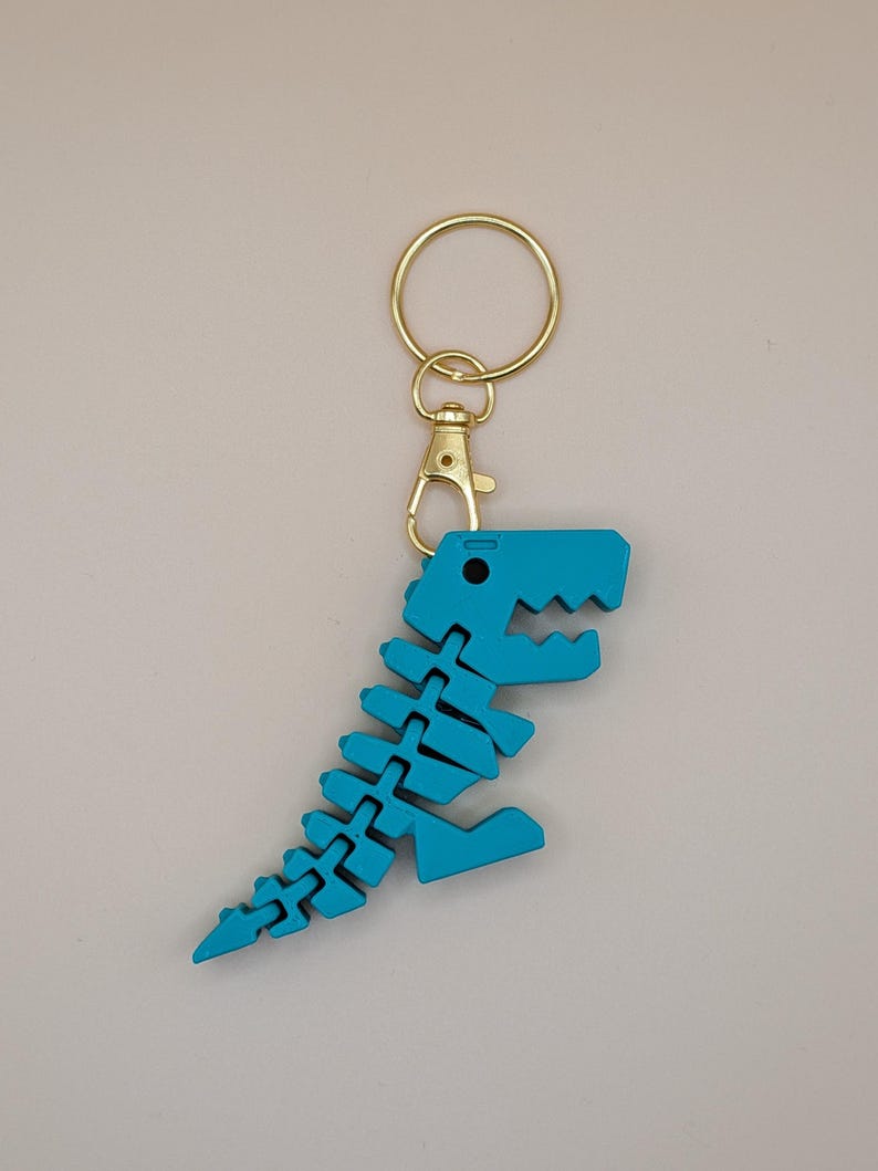May include: A turquoise dinosaur skeleton keychain with a gold-colored clasp and keyring. The dinosaur is made of interlocking pieces, creating a flexible design. The keychain is a fun accessory.