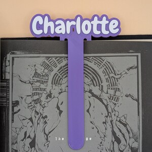May include: A purple 3D-printed bookmark featuring the name Charlotte in white letters with a purple outline. The bookmark has a long, flat, vertical stem extending downwards from the name, which is positioned at the top of a black and white book.