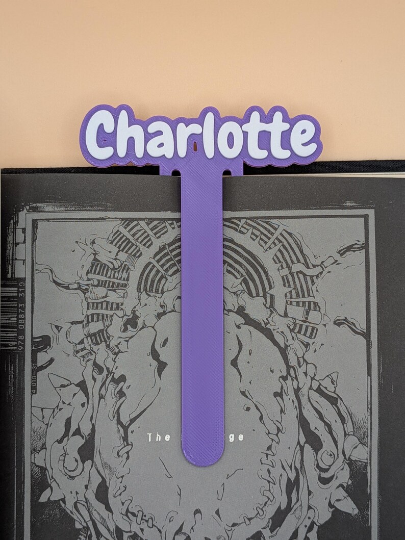 May include: A purple 3D-printed bookmark featuring the name Charlotte in white letters with a purple outline. The bookmark has a long, flat, vertical stem extending downwards from the name, which is positioned at the top of a black and white book.