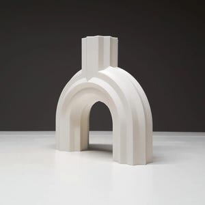 May include: A white, architectural-style decorative object with an archway design. The sculpture has a fluted, textured surface and a rectangular top. The object is displayed on a white surface against a dark gray background.