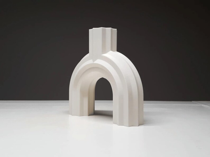 May include: A white, architectural-style decorative object with an archway design. The sculpture has a fluted, textured surface and a rectangular top. The object is displayed on a white surface against a dark gray background.
