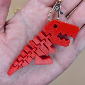 May include: A red 3D-printed dinosaur keychain with a lobster clasp and a gold-colored keyring. The dinosaur is a stylized T-Rex with a segmented body and head. The keychain is held in a hand.