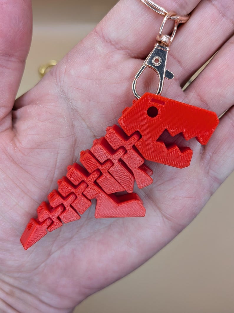 May include: A red 3D-printed dinosaur keychain with a lobster clasp and a gold-colored keyring. The dinosaur is a stylized T-Rex with a segmented body and head. The keychain is held in a hand.