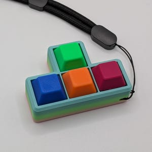 May include: A colorful, Tetris-shaped keychain featuring four square keycaps in blue, orange, green, and magenta. The keycaps are set in a light blue and green gradient frame. A black lanyard is attached for easy carrying.