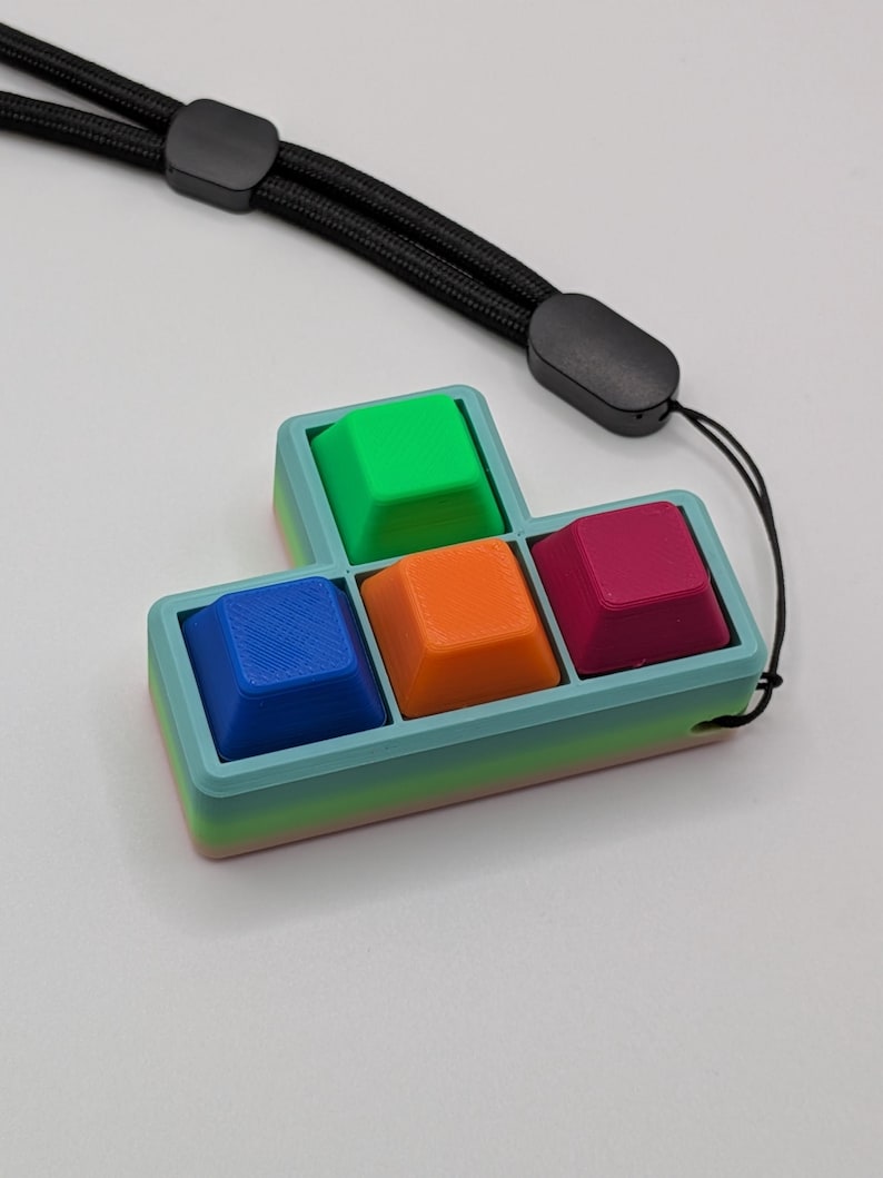 May include: A colorful, Tetris-shaped keychain featuring four square keycaps in blue, orange, green, and magenta. The keycaps are set in a light blue and green gradient frame. A black lanyard is attached for easy carrying.
