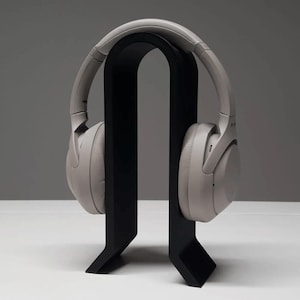 K&ouml;nnte beinhalten: Hellgraue Over-Ear-Kopfh&ouml;rer auf einem schwarzen Kopfh&ouml;rerst&auml;nder. Der St&auml;nder hat ein minimalistisches Design mit einer geschwungenen Oberseite und einem rechteckigen Sockel. Die Kopfh&ouml;rer sind neutralfarben.