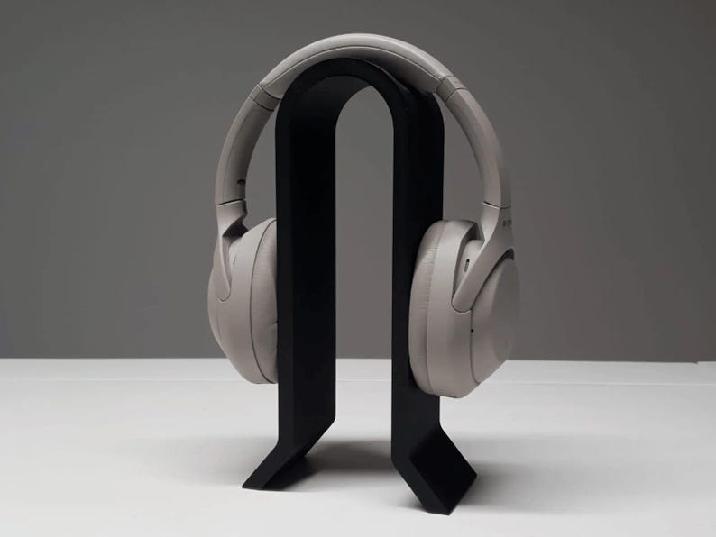 K&ouml;nnte beinhalten: Hellgraue Over-Ear-Kopfh&ouml;rer auf einem schwarzen Kopfh&ouml;rerst&auml;nder. Der St&auml;nder hat ein minimalistisches Design mit einer geschwungenen Oberseite und einem rechteckigen Sockel. Die Kopfh&ouml;rer sind neutralfarben.