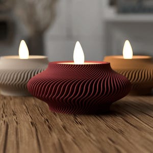 May include: Three decorative candle holders with a wavy, textured design. The candle holders are in cream, burgundy, and tan colors, each with a glowing, flame-shaped light. The candle holders are on a wooden surface.
