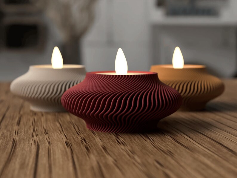 May include: Three decorative candle holders with a wavy, textured design. The candle holders are in cream, burgundy, and tan colors, each with a glowing, flame-shaped light. The candle holders are on a wooden surface.