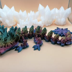 May include: A collection of 3D-printed crystal formations in various sizes and colors. The foreground features a multi-colored, segmented crystal structure with blue, purple, and green hues. Several white crystal clusters are in the background.