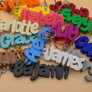 May include: A collection of colorful name keychains in various colors, including pink, blue, green, and white. Each keychain features a different name in a playful, cursive font, attached to a metal keyring. The keychains are a personalized accessory.