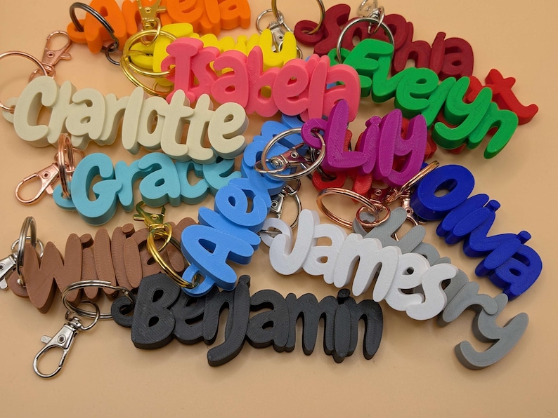 May include: A collection of colorful name keychains in various colors, including pink, blue, green, and white. Each keychain features a different name in a playful, cursive font, attached to a metal keyring. The keychains are a personalized accessory.