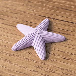 May include: A light purple starfish with textured, ridged arms. The starfish is centered on a wooden surface with visible wood grain. The starfish is a decorative item, possibly for coastal or nautical themed decor.