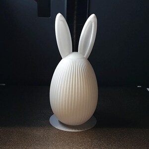 Puede incluir: Una figura de conejo de Pascua blanca, impresa en 3D. El conejo tiene orejas largas y erguidas y un cuerpo en forma de huevo con costillas verticales. Se apoya sobre una peque&ntilde;a base redonda. El fondo es oscuro.