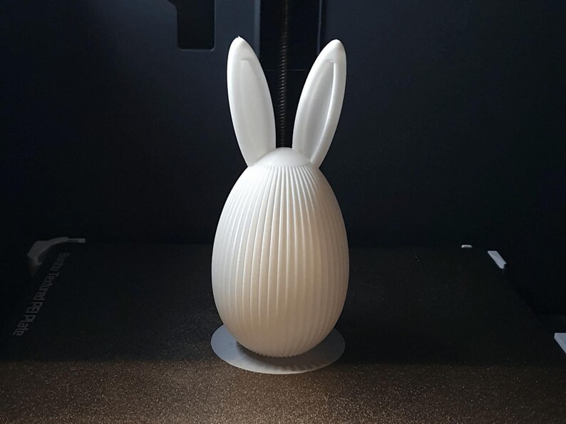 Puede incluir: Una figura de conejo de Pascua blanca, impresa en 3D. El conejo tiene orejas largas y erguidas y un cuerpo en forma de huevo con costillas verticales. Se apoya sobre una peque&ntilde;a base redonda. El fondo es oscuro.