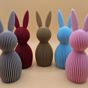 K&ouml;nnte beinhalten: Eine Sammlung von sechs dekorativen Hasenfiguren in verschiedenen Farben. Die Hasen sind vertikal gestreift und haben lange Ohren. Zu den Farben geh&ouml;ren Grau, Burgunderrot, Beige, Blau und Rosa. Die Figuren sind etwa 15 cm hoch.