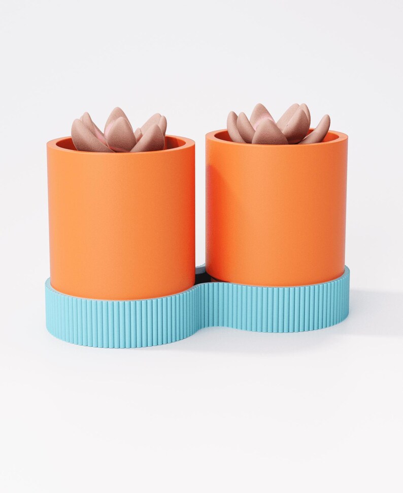 May include: Two orange cylindrical planters with light pink succulent plants, set on a wavy, light blue base. The planters have a smooth finish, while the base features vertical ribbed detailing. The background is plain white.