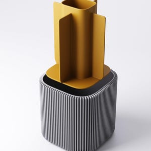 May include: A contemporary desk tidy in two colours. The base is a grey, vertically-lined cube, with a mustard-yellow container on top, featuring four vertical dividers. The background is white.