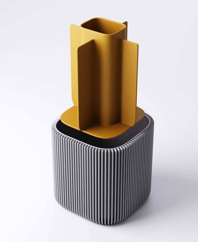May include: A contemporary desk tidy in two colours. The base is a grey, vertically-lined cube, with a mustard-yellow container on top, featuring four vertical dividers. The background is white.